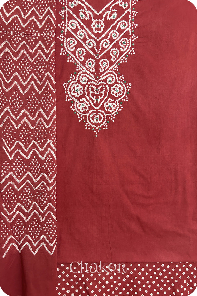 A brick red kurta set fabric with intricate bandhani & mirror embroidered yoke, paired with a brick red bottom & odhani placed on the left side featuring a zigzag design.