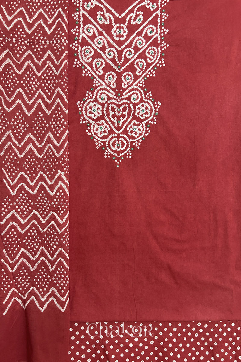 A brick red kurta set fabric with intricate bandhani & mirror embroidered yoke, paired with a brick red bottom & odhani placed on the left side featuring a zigzag design.