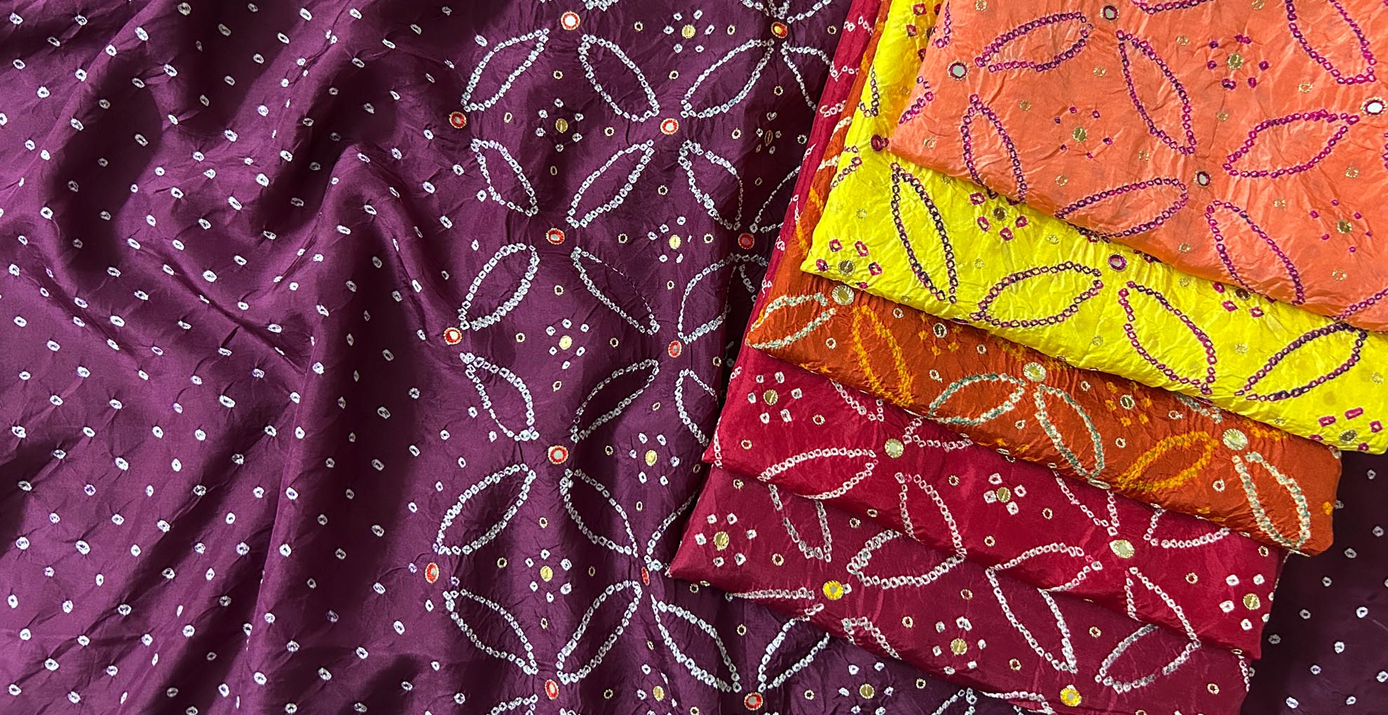 Stack of colourful bandhani mulberry silk kurta fabric with rai bandhej patterns adorned with mirror work, sequins and zari embroidery.