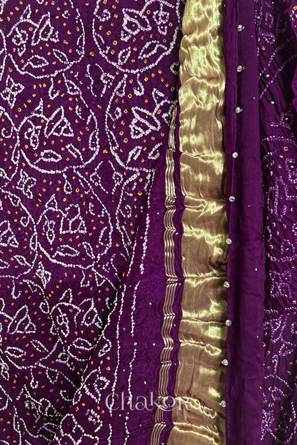 Close-up of a rai bandhej dupatta in magenta featuring white & yellow bandhani dots along with mukaish embroidery & shimmering gold band on pallu.Text reads Chakor brand watermark.

