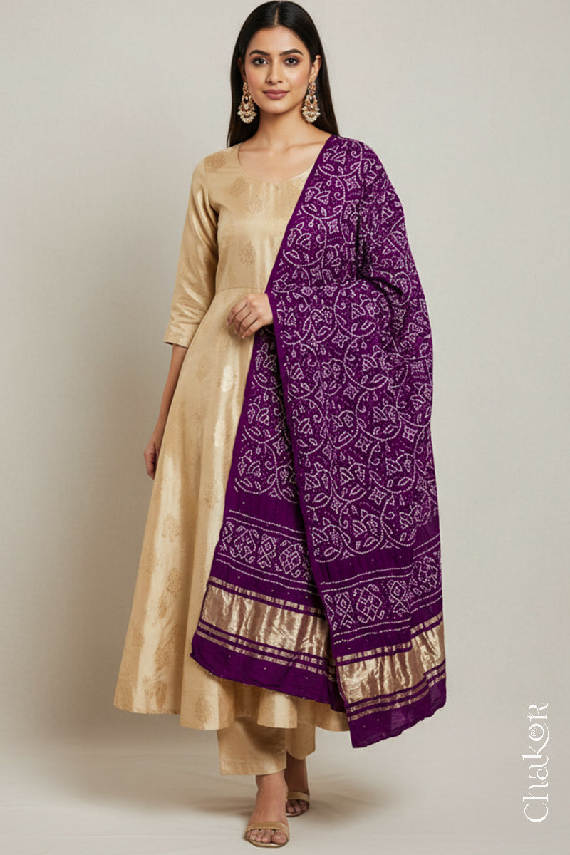 A woman in a beige ethnic outfit, wearing a colorful rai bandhej dupatta in magenta with golden zari pallu, stands gracefully against a light background.Text reads Chakor brand watermark.
