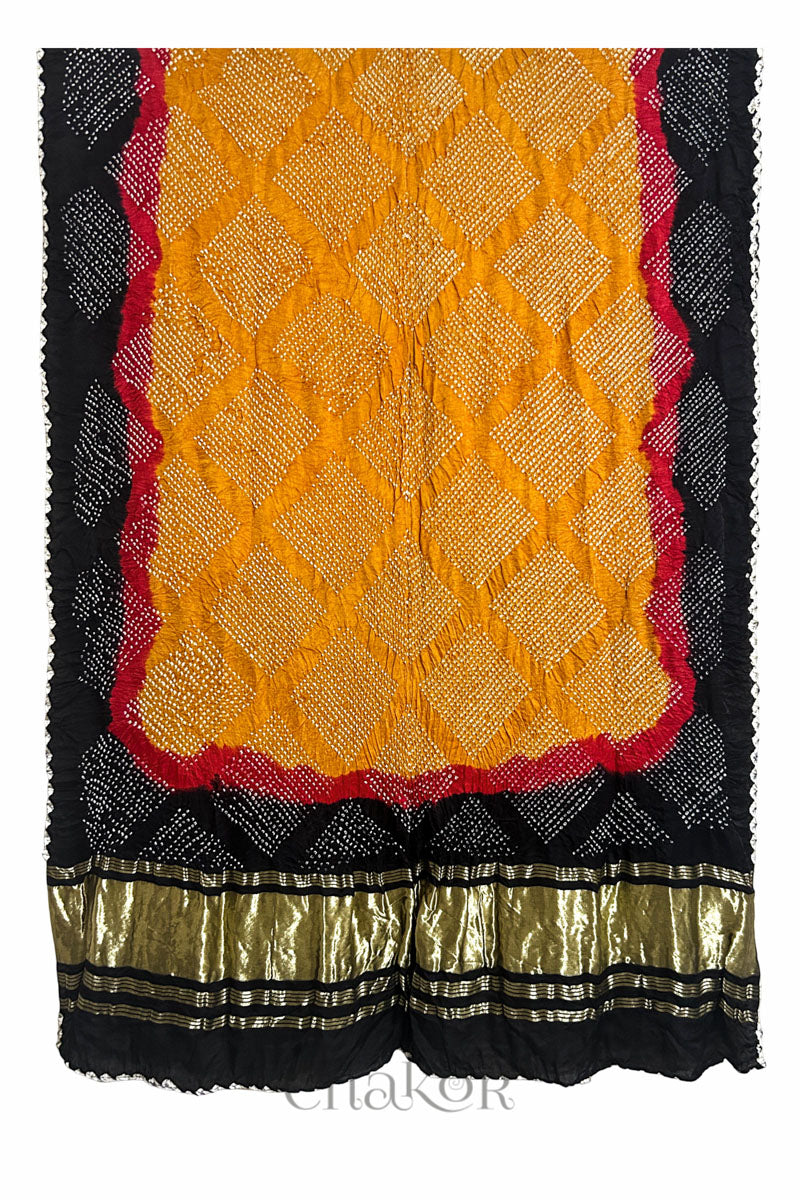 A vibrant dupatta showcasing intricate rai bandhej on mustard yellow, red accents, and black border with a shimmering gold band on the pallu and Chakor brand watermark.