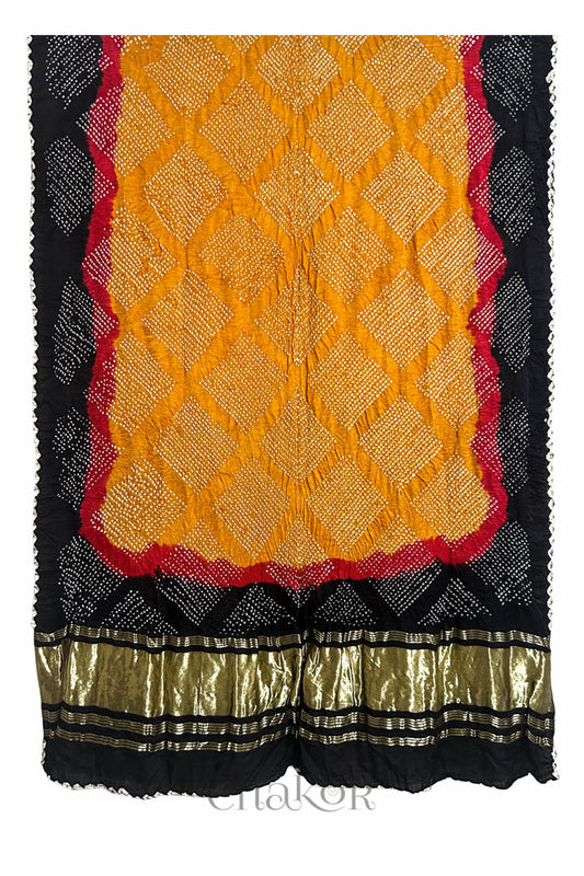 A vibrant dupatta showcasing intricate rai bandhej on mustard yellow, red accents, and black border with a shimmering gold band on the pallu and Chakor brand watermark.
