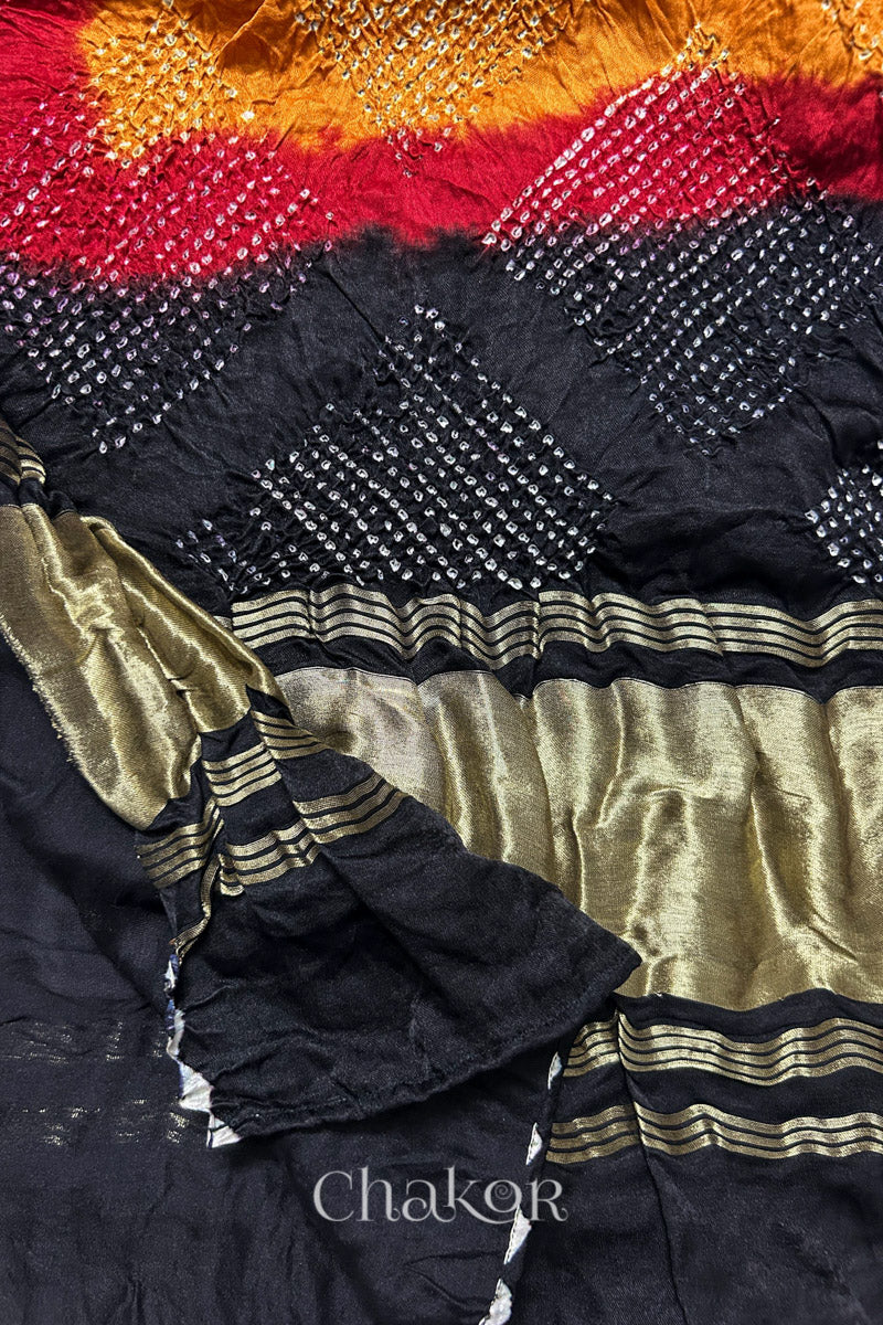 A colourful dupatta showcasing rai bandhej on mustard yellow, red and black pallu with tissue band folded on corner. Text reads Chakor brand watermark.