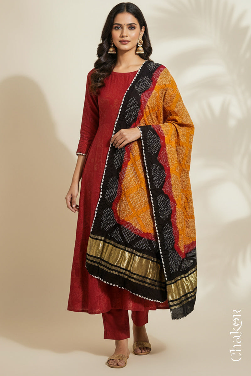 A woman in a red ethnic outfit, wearing a colourful rai bandhej dupatta with in mustard yellow, red and black colour, stands gracefully against a light background.