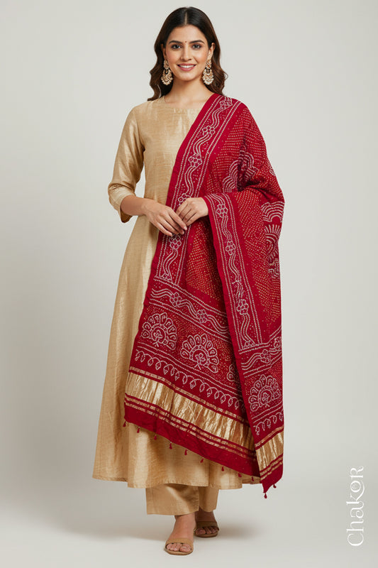 A woman in a beige ethnic outfit, wearing a colorful rai bandhej dupatta in red with golden zari pallu & tassels, stands gracefully against a light background.Text reads Chakor brand watermark.
