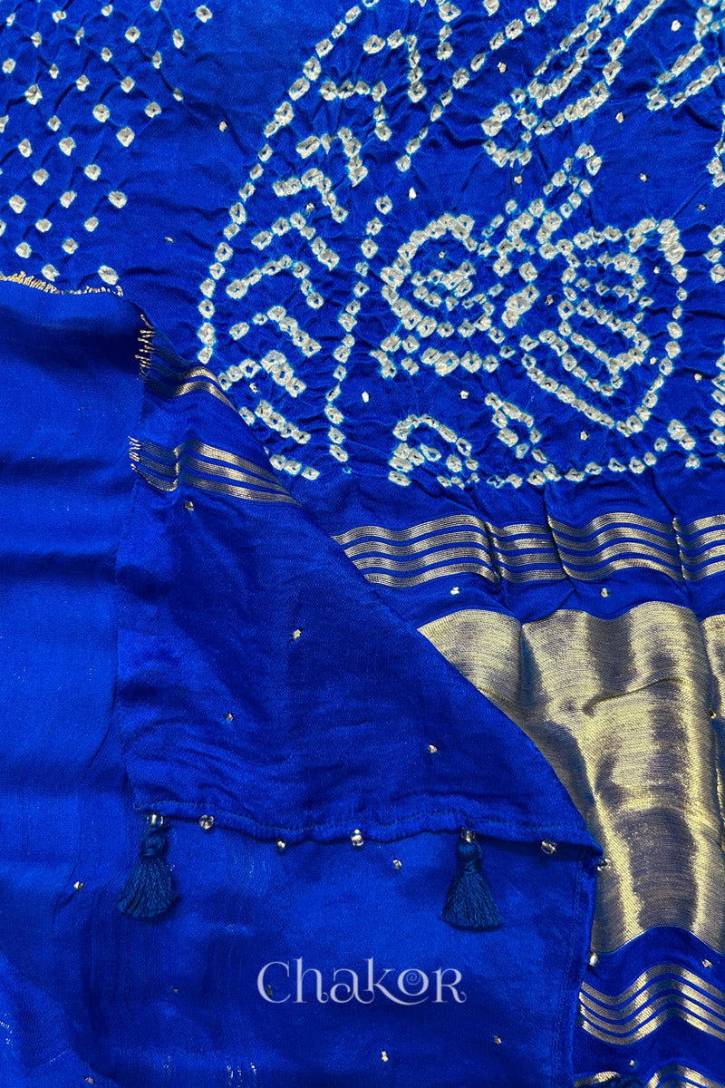 A colourful dupatta showcasing rai bandhej in blue ombre dyed accented with mukaish work with tissue band folded on corner. Text reads Chakor brand watermark.
