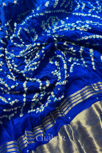 A colourful dupatta showcasing rai bandhej in blue ombre dyed with golden tissue band placed in swirling style. Text reads Chakor brand watermark.

