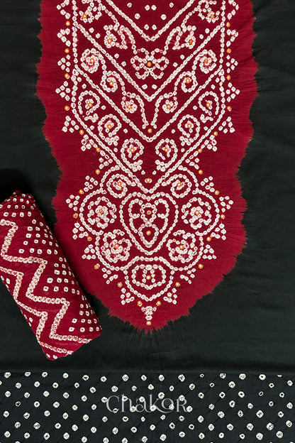A classic Black set fabric featuring mirror embroidered contrast yoke in red, matching coloured bandhani bottom and red dupatta rolled beautifully and placed on the fabric. Text reads Chakor.