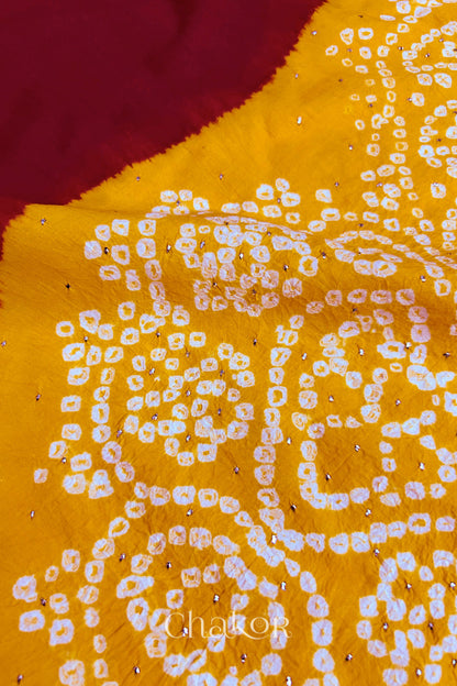 Close-up of red fabric adorned with intricate traditional bandhani patterns and sparkly mukaish embroidery on yellow yoke, showcasing beautiful craftsmanship.
