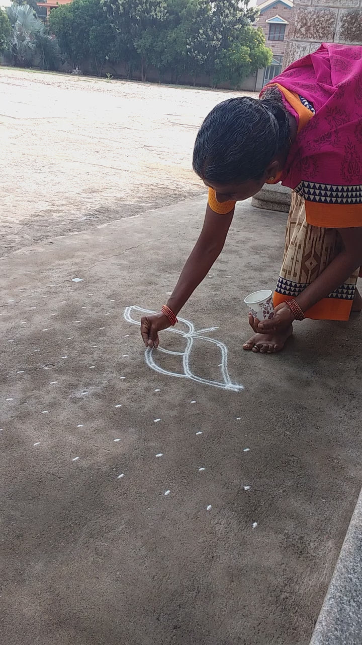 Load video: A woman in a simple saree crouches at the entrance of a craft exhibit, gently arranging a rangoli design with care. Her quiet gestures speak of warmth and intention, captured in a soft, reflective tribute to unseen acts of kindness.